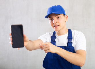 Handyman in blue jumpsuit and cap shows dark empty mobile phone screen and gestures with his hand in bewilderment.Studio, grey background