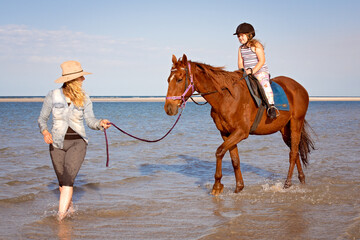 Young woman leading brown horse with young girl riding, through shallow ocean water at beach