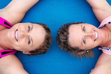 Two young women in fitness gear lying on blue exercise mat, looking straight up at camera, smiling, overhead view