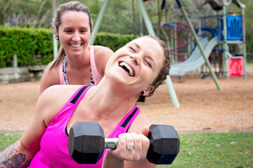 Two women laughing while exercising with dumbbells outdoors in park