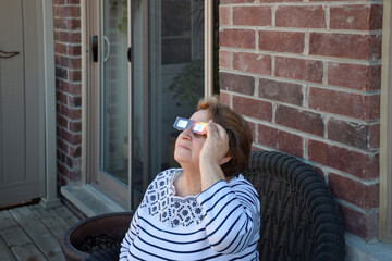 Senior woman sitting on a balcony alone watching solar eclipse using special protection eyeglasses - obrazy, fototapety, plakaty