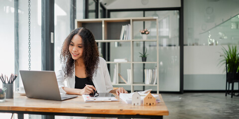 Smiling real estate agent engaged in work on her laptop at a wooden desk, surrounded by documents...