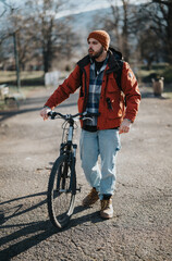 Casual bearded man in a beanie walking with a mountain bike on a sunny day, showcasing outdoor leisure activity.