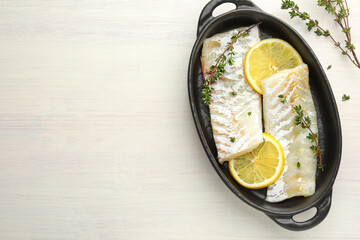 Fresh raw cod fillets with thyme and lemon in baking dish on white wooden table, top view. Space for text