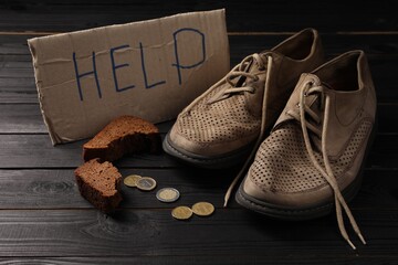 Poverty. Cardboard sign with word Help, old shoes, pieces of bread and coins on black wooden table