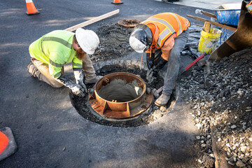 Construction workers installing a sewar access point collar in the middle of a newly paved road
