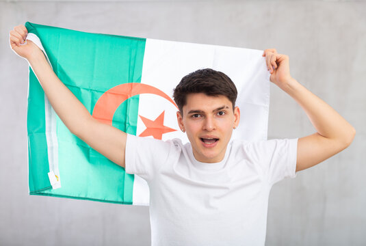 Happy young man with flag of Algeria in hands posing against light unicolored background