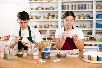 Concentrated young man and woman in aprons engraving on clay cup with cutter in pottery workshop