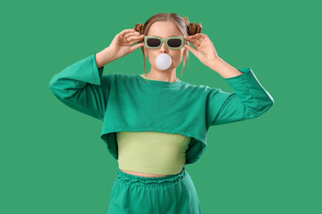 Beautiful young woman in stylish outfit chewing gum on green background