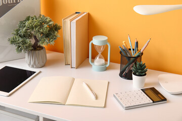 Comfortable workplace with tablet computer, hourglass and notebooks near orange wall