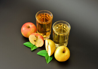 Two glasses of fruit juice on a black background, next to pieces of a red and yellow apple with leaves.