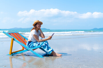 Happy Asian woman enjoy outdoor lifestyle travel ocean on summer holiday vacation. Attractive girl relaxing and using smartphone for online shopping or chatting at tropical island beach in sunny day.