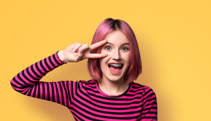 Photo image - happy pink beautiful woman in braces brackets showing v-sign victory peace hand gesture two fingers, opened mouth, isolate yellow wall background. Girl at studio. Dental care ad concept