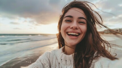 A woman joyfully stands on a sandy beach next to the ocean, capturing a selfie