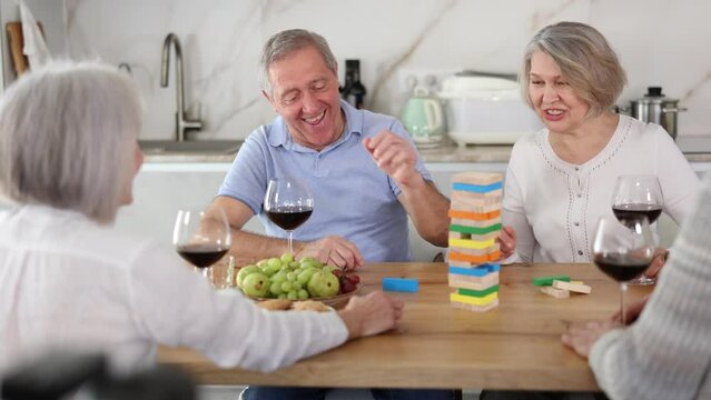 Elderly happy people playing Jenga at home, woman is removing a wooden block from the tower and the others are watching