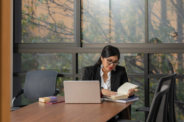 a business woman reading a book in the office during a work day, sitting at her desk with her laptop