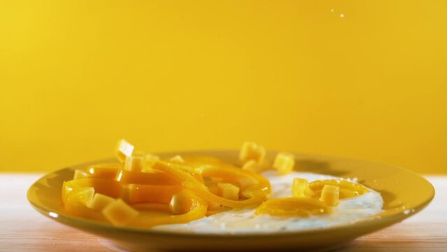 In a slow-motion sequence, salts and vegetable pieces gracefully descend onto a plate with two eggs, set against a yellow backdrop, depicting the art of food preparation and a breakfast scene.