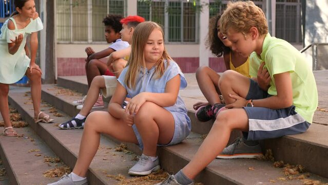 Group of modern positive tweenagers hanging out on streets of city on warm summer day, friendly blabbing while sitting on steps outdoors