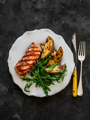 Delicious lunch - grilled salmon, baked potatoes and arugula salad on a dark background, top view
