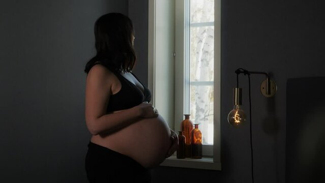 Heavily pregnant woman touches bump and gazes out window