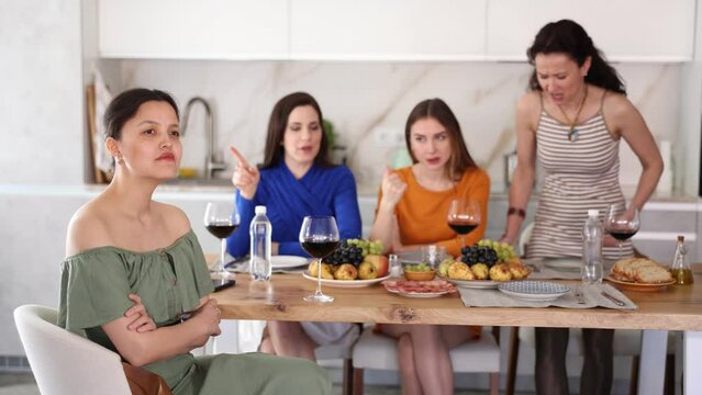 Upset young Asian woman sitting with crossed arms at table during home gathering while female friends expressing discontent and criticizing in background