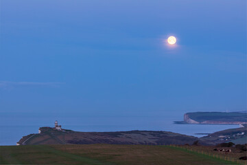 Full moon setting over Seaford Head and Belle Tout lighthouse on the cliff edge of Beachy Head east...