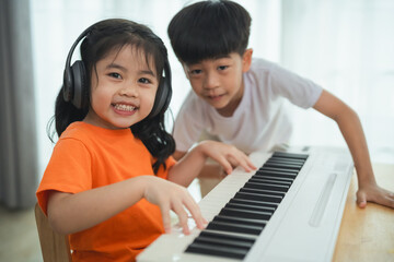 Two children are playing the piano. One is wearing an orange shirt. They are smiling and seem to be enjoying themselves