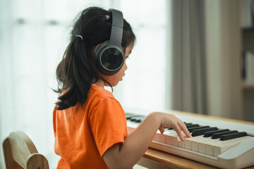 A young girl is sitting at a piano, wearing headphones. She is wearing an orange shirt. The room is...