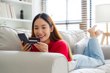 Young asian woman lying on sofa in living room, makes online banking payments through the internet from bank card on cell phone. Shopping online on mobile with credit card - obrazy, fototapety, plakaty