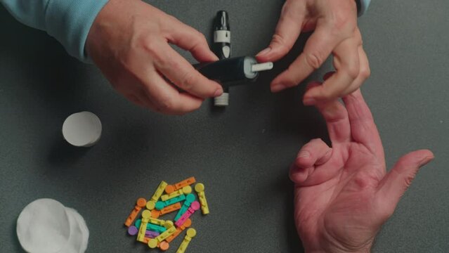 An elderly couple sitting at a table. A wife helps her diabetic husband do a quick blood sugar test using a portable glucometer with replaceable needles. Hands close up