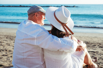 Rear view of bonding senior couple hugging sitting on the beach face the sea enjoying the sunset in vacation or retirement