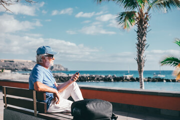 Senior bearded man holding smartphone sitting outdoors on bench face the sea enjoying sunny day,...
