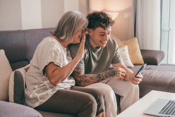 Video call concept. Happy senior retired woman sitting on sofa with young nephew waving hands video calling by mobile phone using technology for online webcam connection with distant family or friends - obrazy, fototapety, plakaty
