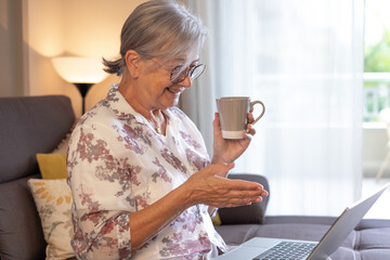 Attractive Caucasian senior woman sitting on sofa at home watching funny movie or video on laptop while drinking cup of coffee