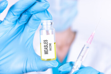 Text MEASLES VACCINE of is written on a bottle with the background of a doctor with a syringe in a medical glove and mask. Medical concept.