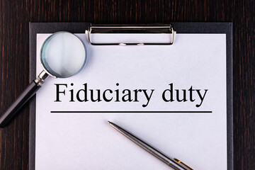 Text FIDUCIARY DUTY is written on a notebook with a pen and a magnifying glass lying on the table....