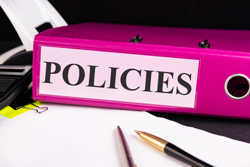 Text POLICIES is written on a folder lying on a stack of papers with a pen on the table. Business...