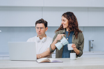 A woman pours coffee for her partner who is focused on work at the laptop, exemplifying a...