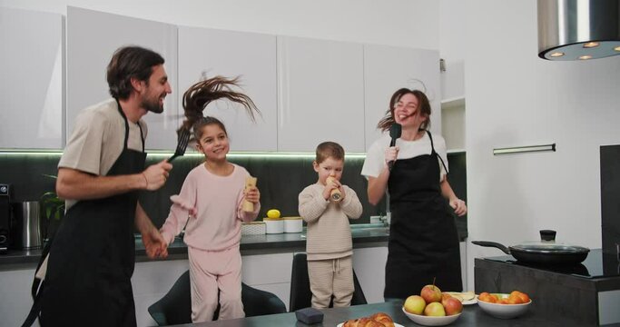 Happy family brunette man with stubble in a black apron together with his brunette wife and two small children dancing and having fun in a modern kitchen before starting breakfast in a modern