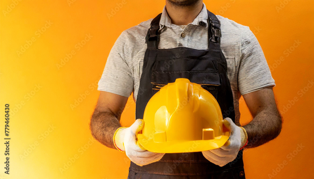 Poster manual worker with protective work gloves holding a yellow safety helmet on a yellow and orange back