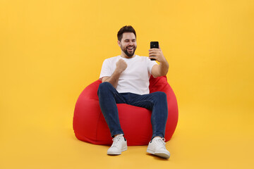 Happy young man using smartphone on bean bag chair against yellow background
