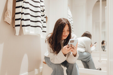 Korean teenage girl takes a selfie in new clothes in the mirror and sends it to her friend via phone in a shopping mall. Retail and consumerism. Sale promotion and shopping concept. Part of a series - 779467441