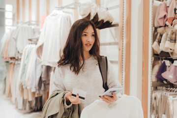Korean teenage girl holds a credit card in her hands and wants to pay for new clothes in a shopping mall. Retail and consumerism. Sale promotion and shopping concept. Part of a series - 779472049
