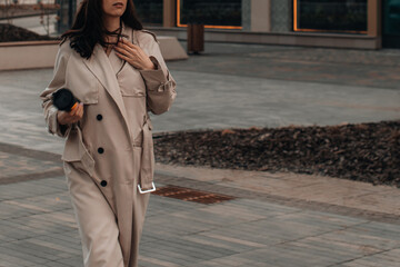 Woman wearing beige long seasonal trench coat, hand in pocket, walking in the city. Street style photo shoot