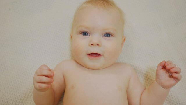 Blue eyed charming baby boy lies on his back, looks at the camera and smiles. Portrait of a cute little child with blond hair. Close-up of the face of a white Caucasian baby.