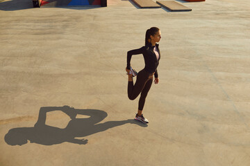 Young woman in sports clothes having outdoor fitness workout on city street, standing on grey concrete sportsground surface, getting ready to start jogging and doing warming up leg stretching exercise