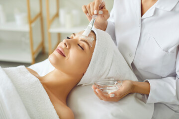 Cosmetologist applying moisturizing mask with brush to a young relaxed smiling young woman in spa...