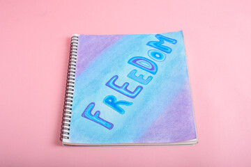 Notebook with the word freedom drawn with colored chalk. Painted notebook on pink background.