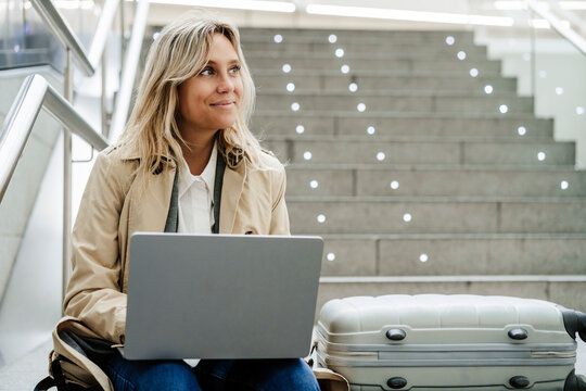 Smiling freelancer with laptop sitting by luggage on steps at train station