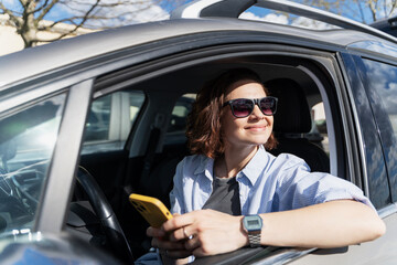 Smiling Caucasian woman in sunglasses sitting in a car in the parking lot driver's seat looking at the smartphone